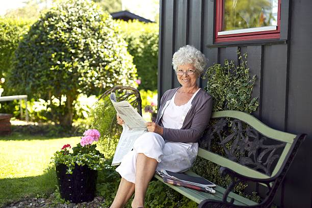 Relaxed elder woman sitting on a bench in backyard garden reading a newspaper looking at camera and smiling
