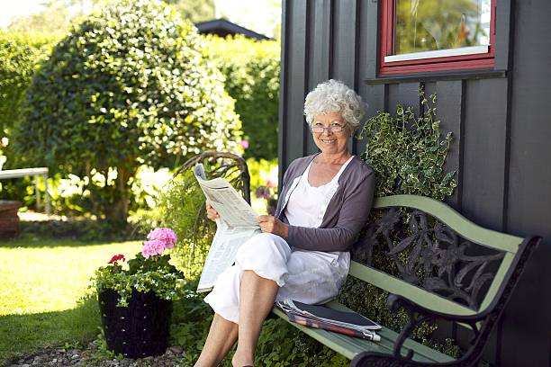Elder woman reading newspaper in backyard garden Relaxed elder woman sitting on a bench in backyard garden reading a newspaper looking at camera and smiling sitting on bench stock pictures, royalty-free photos & images