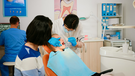 Stomatologist holding sterilized dental tools talking with little patient before stomatological intervation. Nurse and doctor wearing masks cleaning teeth of child lying on chair with open mouth