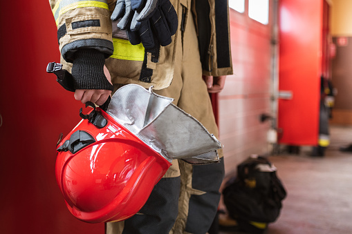 Back view of crop anonymous firefighter in protective uniform with yellow lines standing with long hose dripping water on pavement in daylight. Risk professions concept