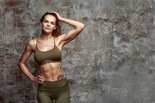 Shot of good looking sporty slim woman dressed in sportsclothes with muscular abs has active lifestyle, poses against grey concrete background. Lifestyle concept