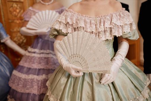 Closeup of ladies wearing big ballgowns holding fans while waiting for partners during ball in palace, copy space