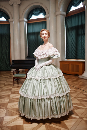 Vertical full length portrait of beautiful young woman wearing classic ballgown standing in spotlight in palace hall