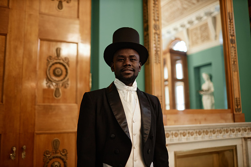 Waist up portrait of smiling Black gentleman wearing top hat looking at camera in palace
