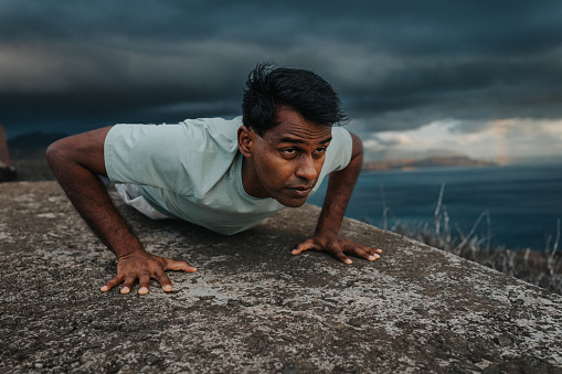 An active Indian man holds a lot plank position while exercising at the summit of a mountain hike overlooking the ocean in Hawaii on a cloudy morning.