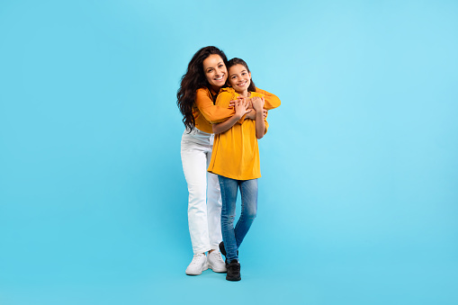 Happy european mother hugging her lovely daughter, standing over blue studio background, full length shot, free space. Young mom and he child girl bonding and smiling to camera