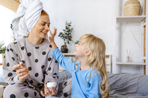 Smiling mother and her little cute daughter in pajamas enjoying in bed. Their hair is wrapped in towels