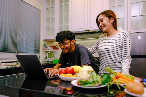 Happy asian couple cooking together while using laptop in the kitchen. Circular habits in the kitchen.