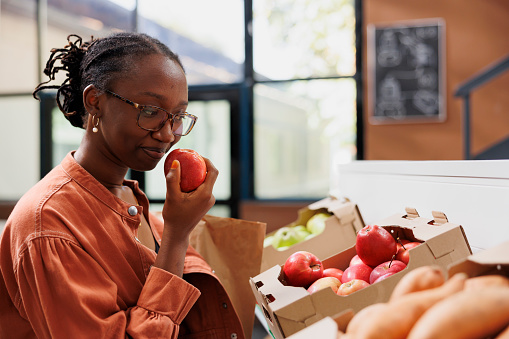 African american customer grasping and smelling fresh red apple, choosing best product in eco friendly supermarket. Black lady picking locally grown produce from boxes on shelves in convenience store.