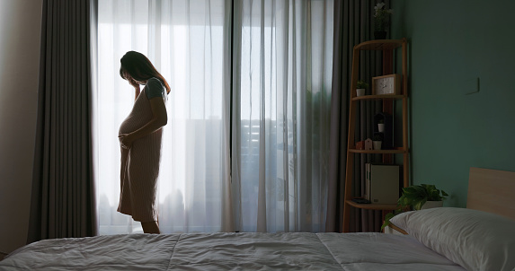 silhouette asian pregnant woman feel depressed about being new mom while standing by windows