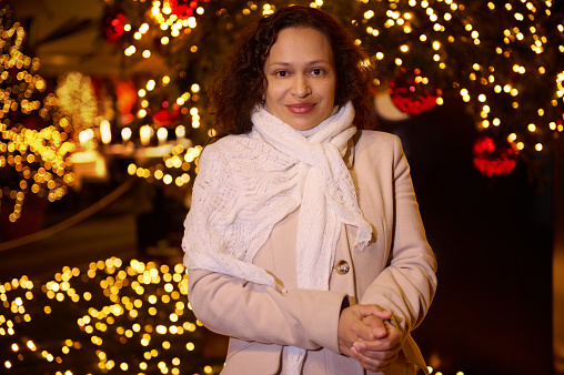 Portrait of a beautiful woman in warm clothes, smiling looking at camera, standing against illuminated street background. Enjoying the upcoming winter and Christmas holidays. December Xmas fairground