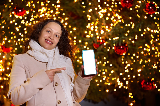 Beautiful African American woman showing smart phone with white blank digital screen, with copy space for ad or mobile app, standing against a Christmas tree decorated with holiday lights and garlands