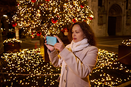 Beautiful multi ethnic curly haired brunette woman, holding smart mobile phone, photographing the Christmas fairground in the city square illuminated by holiday lights and garlands at night time