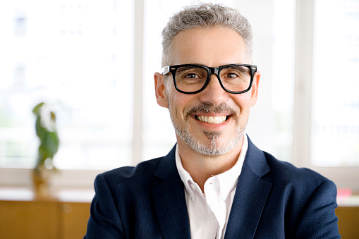 Close-up portrait of cheerful and serene mature modern businessman in smart casual wear, stylish glasses, headshot of senior 60s gray-haired male manager, leader looking at camera with toothy smile