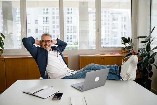 Smiling senior businessman sitting at the desk, putting his feet on the table, holding hands behind head, relaxing after a productive day, self-assured, works in a comfortable and modern office