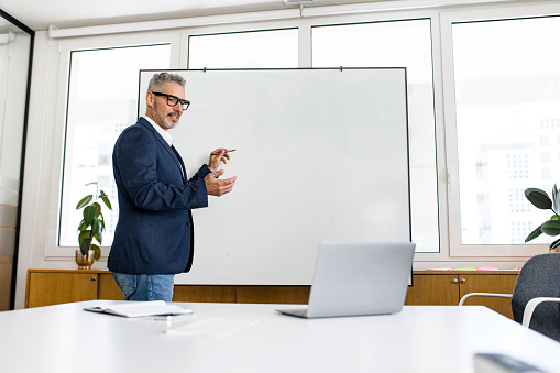 Confident mature businessman conducting webinar, leading business training online. Online coach stands near flip chart in front of laptop and explains something to online audience