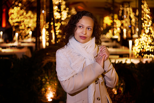 Attractive curly haired woman in stylish warm coat, holding hands cupped, dreamily looking aside, making cherished wish for Christmas, standing on the street with beautiful lightings on the background