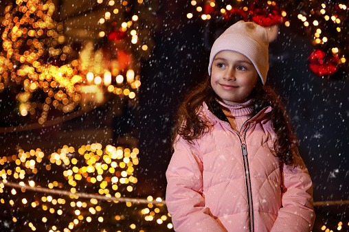 Confident portrait of adorable little kid girl smiling dreamily looking away, standing outdoor against lightened street and Christmas tree at funfair. People, holidays and festive life events concept