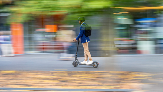 Businessman riding electric scooter in urban downtown district street, side wide shot with blurred background