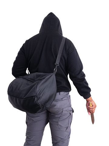 An unidentified male criminal wearing a black hoodie and covering his face, holding a sharp knife and carrying a large black money bag. On a white background with a clipping path.