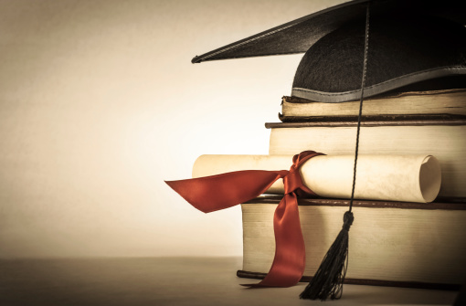 A mortarboard and graduation scroll, tied with red ribbon, on a stack of old battered book with empty space to the left.  Slightly undersaturated with vignette for vintage effect.