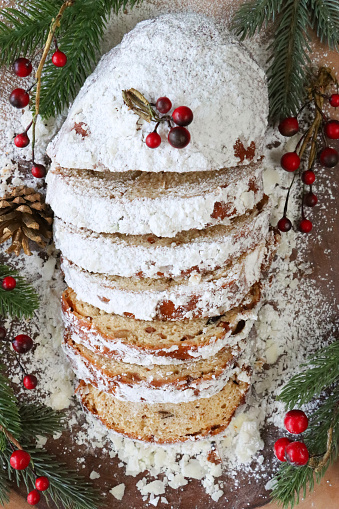 Stock photo showing close-up, elevated view of a sliced Christmas stollen cake, made with with marzipan, currants, raisins and decorated with powdered icing sugar surrounded by Spruce needles, pine cone and red berry decorations.