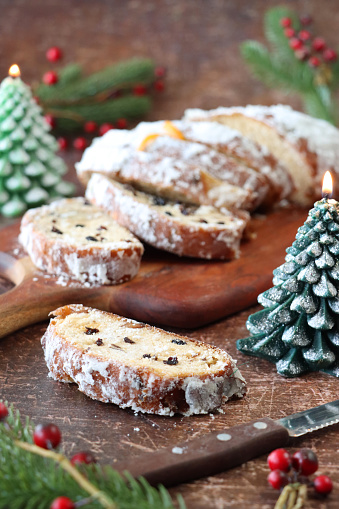 Stock photo showing close-up view of wooden chopping board containing  a sliced Christmas stollen cake, made with with marzipan, currants, raisins and decorated with powdered icing sugar surrounded by burning Christmas tree shaped candle, Spruce needles an red berry decorations.