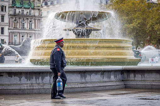 Trafalgar Square, London, England - October 30th 2023:  Man in a uniform walking with a bottle of water in front of one the famous fountains