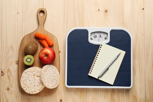 Healthy diet. Scale with notebook and products on wooden table, flat lay