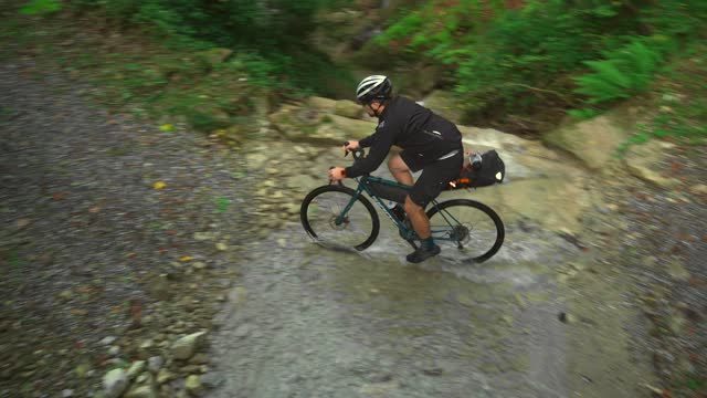 Male cyclist crossing river in mountains on gravel bicycle. Bicycle tourist bikepacker crosses mountain river in the forest in autumn. Bike adventure. Bicyclist traverses mountain stream on cycle.