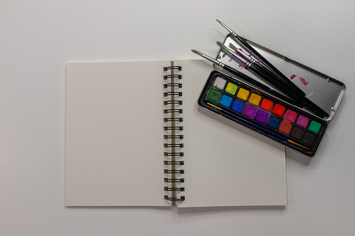 Flat lay composition of open sketch book with watercolor palette on white background with copy space