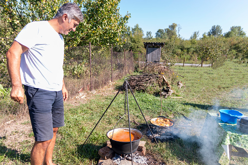 Mature man cooking food on campfire outdoors. Delicious meal on barbecue grill with coal outdoors picnic. Assortment of fresh healthy food on a BBQ grilling over a hot fire in a green grassy spring or summer field