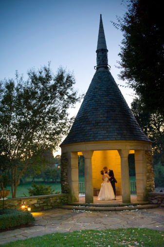 Picture perfect of bride and groom kissing at sunset in a garden's stone gazebo. Vertical portrait size. 