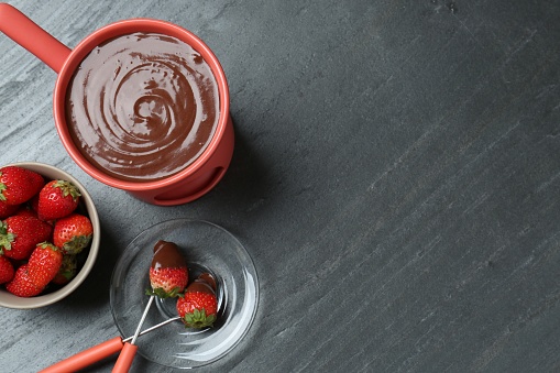 Fondue pot with melted chocolate, fresh strawberries and forks on grey table, flat lay. Space for text