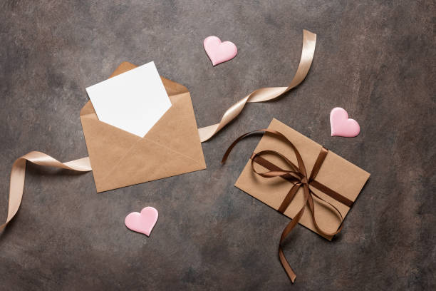 istock Open brown envelope flat lay with blank card mockup decorated with silk ribbon and silk hearts on dark vintage background. Valentine's Day card. Top view. 1855471110