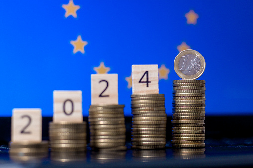 Number 2024 representing new year next to one euro coin on highest stack of coins in front of European Union flag illustrating financial politics in 2024 or inflation