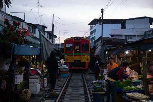 A full sized train travels through a busy market space. The Rom Hub Market is famous for an active railroad passing directly through the busy outdoor market space in Mae Klong, Thailand