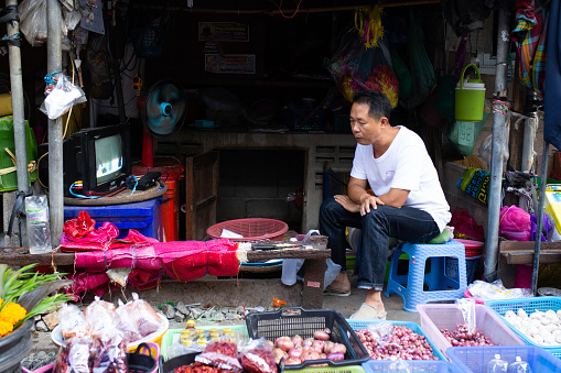 A portrait of a street vendor watching tv in a market place. In the small city of Mae Klong, Thailand