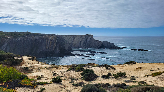 Praia de Almograve beach with ocean waves, cliffs and stones, wet golden sand and green vegetation at wild Rota Vicentina coast, Odemira, Portugal