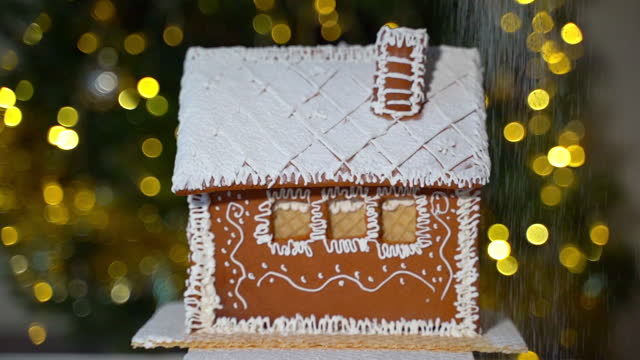 A handmade gingerbread house spins against the backdrop of a Christmas tree with powdered sugar sprinkled on top