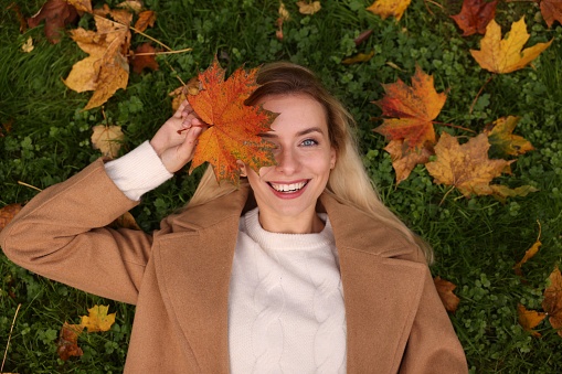 Smiling woman lying on grass and covering eye with autumn leaf, top view