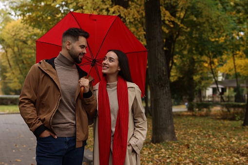 Happy young couple with red umbrella spending time together in autumn park, space for text