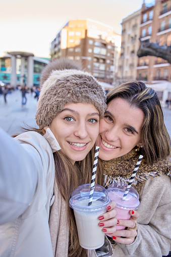vertical photograph of a young female couple taking a selfie with their smartphone holding a smoothie.