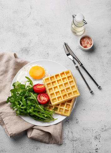 Waffles with fried egg, cherry tomatoes and arugula on a white plate on a gray texture background with spices. The concept of a healthy and tasty breakfast. Top view.