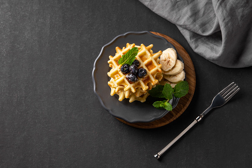 A stack of belgian waffles with kiwi, banana slices and mint, drizzled with honey syrup on a gray plate on a dark background. The concept of a healthy dietary and delicious breakfast. Top view