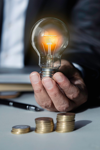 Businessman is holding a light bulb and with a pile of coins on the table.