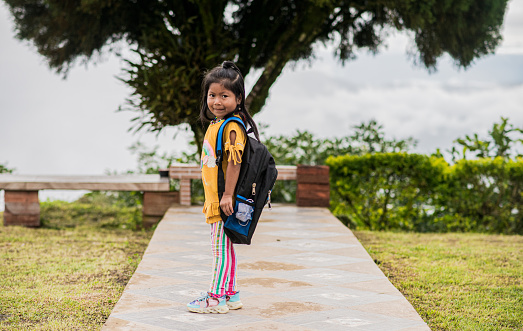 Portrait of a little indigenous girl walking down a hallway and in the background a tree with her backpack on her shoulders.