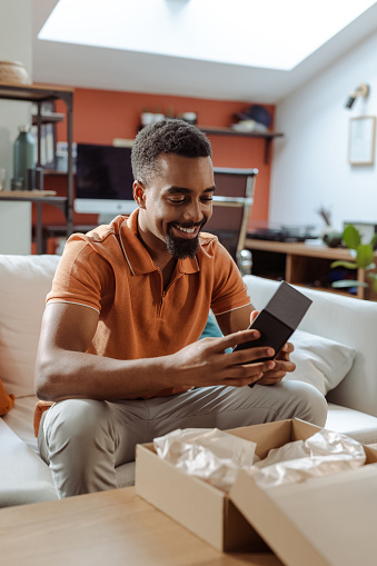 Smiling African American young man opening package with online order and smiling. He is sitting on the sofa in the living room and opening cardboard box