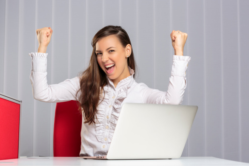 Excited female executive screaming and celebrating with raised clenched fists her business victory in front of laptop in the office.
