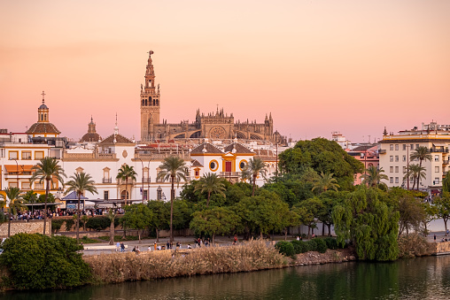 Sunset in Sevilla. Panoramic golden sunset view of La Giralda tower and rooftop of Seville Cathedral rising behind soaring dome of the Church of the Divine Saviour. People enjoying the sunset in the riverside. Seville, Andalusia, Spain